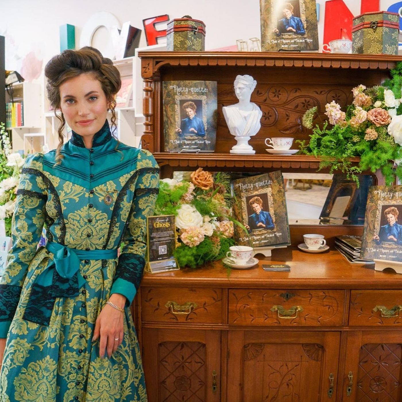 Victorian-costumed host beside the Hetty-quette book display and florals